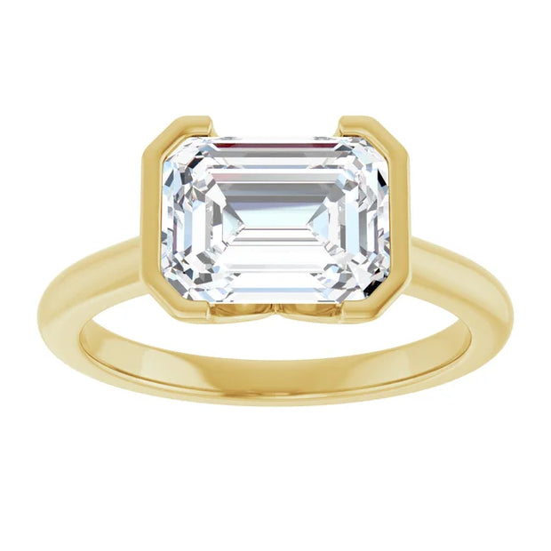 Top view of a 14K yellow gold engagement ring featuring a single, rectangular emerald cut diamond in the center. The diamond is held securely in a delicate half bezel setting that allows light to pass through the top and sides of the stone, revealing its brilliance
