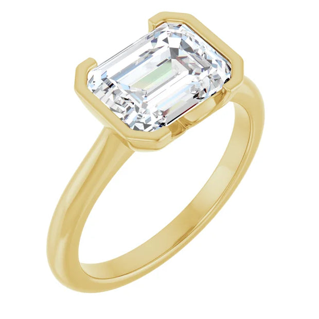 Perspective view of a 14K yellow gold engagement ring, as if worn on a finger. The ring features a gleaming emerald cut diamond held securely in a half bezel setting, revealing the elongated facets of the diamond from a slightly angled view.